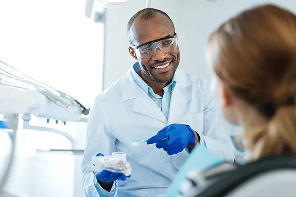 Smiling dentist explaining brushing tips to a patient using a dental model as part of preventive dentistry education.